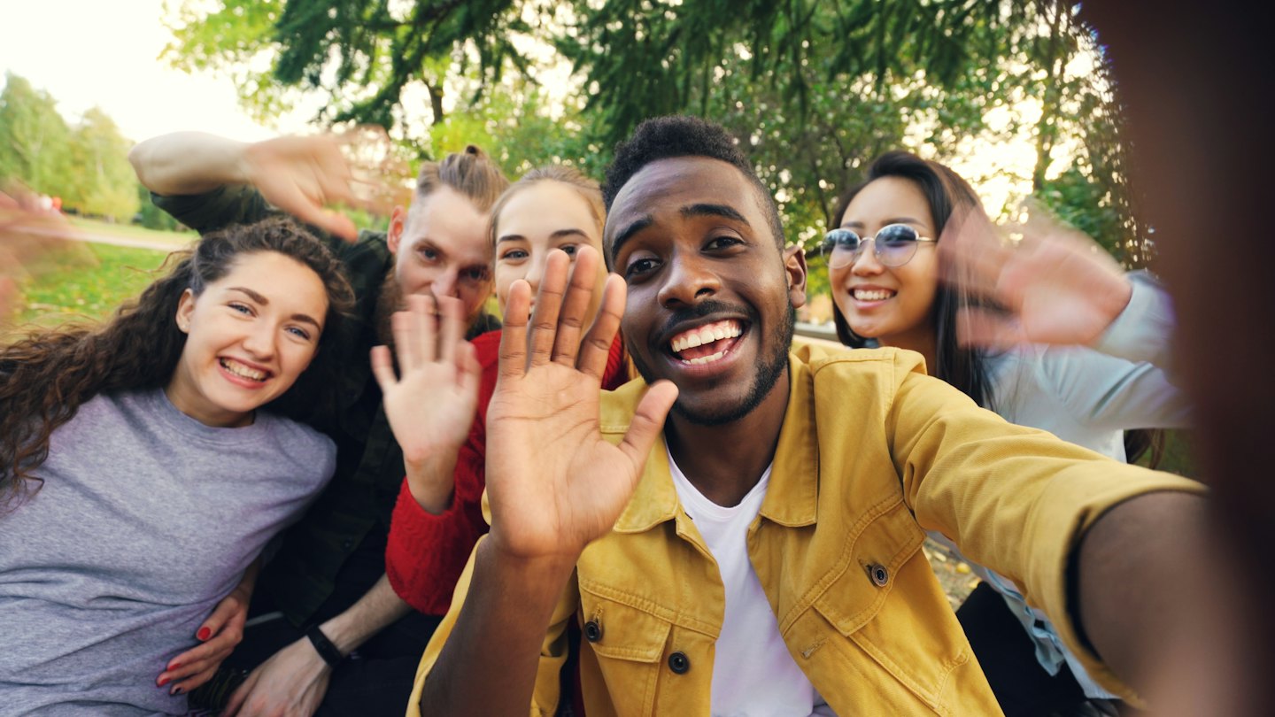Cinque amici sorridono e salutano per un selfie in un parco.